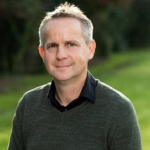 man with black shirt and greay jumper looks at the camera