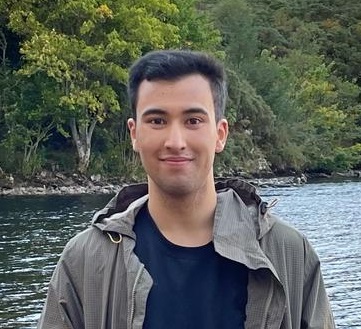 young man with short hair, blue tshirt and grey jacket, infront of a river and river bank on far side