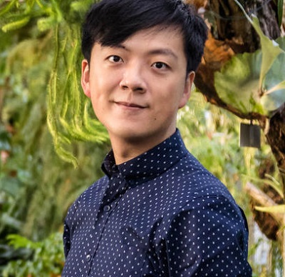 man with short black hair in blue shirt with polka dots standing in front of a tree