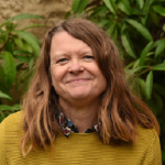 woman with long hair with a blouse and a mustard jumper looks at camera