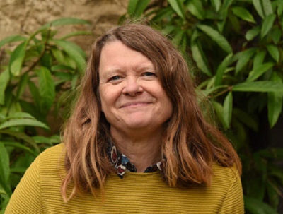 woman with long hair with a blouse and a mustard jumper looks at camera