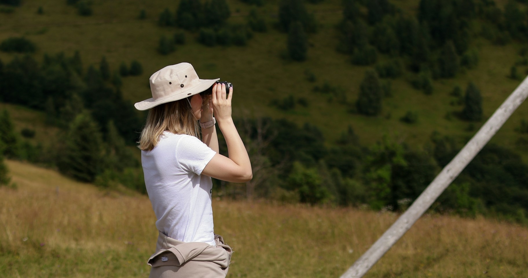 Woman with a hat on looking through binoculars, green hills behind